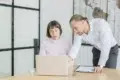 A man in a white shirt leans over a woman seated at a desk to look at her laptop screen in a bright, modern office.
