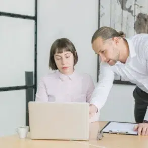 A man in a white shirt leans over a woman seated at a desk to look at her laptop screen in a bright, modern office.