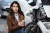 Worried woman holding a smartphone next to a heavily damaged car.