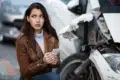 Worried woman holding a smartphone next to a heavily damaged car.