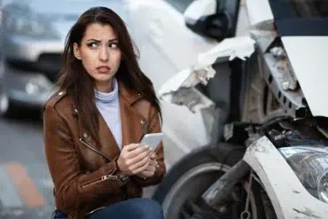 Worried woman holding a smartphone next to a heavily damaged car.