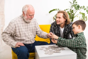 An elderly man, a younger man, and a boy are assembling a jigsaw puzzle at a white table.