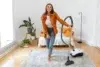 Young woman in jeans and an orange shirt vacuums a patterned rug in a bright, plant-filled room.