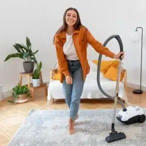 Young woman in jeans and an orange shirt vacuums a patterned rug in a bright, plant-filled room.