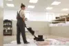 Woman in overalls operates a floor buffer on a rug in a cleaning facility.