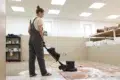 Woman in overalls operates a floor buffer on a rug in a cleaning facility.