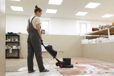 Woman in overalls operates a floor buffer on a rug in a cleaning facility.