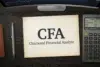 A desk with a paper reading "CFA Chartered Financial Analyst," a calculator, a pen, and a computer monitor displaying a stock chart.