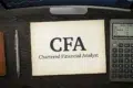 A desk with a paper reading "CFA Chartered Financial Analyst," a calculator, a pen, and a computer monitor displaying a stock chart.
