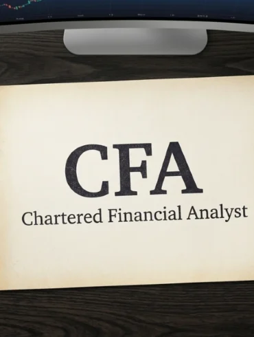 A desk with a paper reading "CFA Chartered Financial Analyst," a calculator, a pen, and a computer monitor displaying a stock chart.