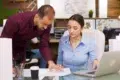 A man in a burgundy shirt points at a document while a woman in a blue shirt looks at it, with a laptop open on the desk.