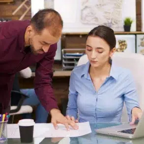 A man in a burgundy shirt points at a document while a woman in a blue shirt looks at it, with a laptop open on the desk.