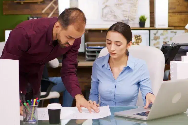 A man in a burgundy shirt points at a document while a woman in a blue shirt looks at it, with a laptop open on the desk.