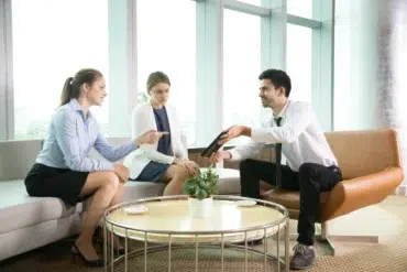 Three business professionals in a modern office lounge discussing a tablet.