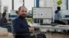Man in a wheelchair smiling at the camera in a modern office with colleagues working in the background.