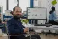 Man in a wheelchair smiling at the camera in a modern office with colleagues working in the background.