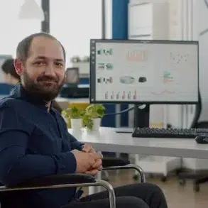 Man in a wheelchair smiling at the camera in a modern office with colleagues working in the background.