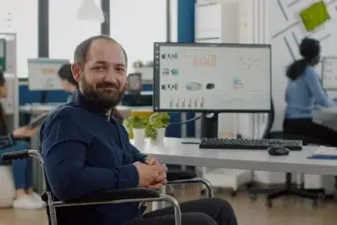 Man in a wheelchair smiling at the camera in a modern office with colleagues working in the background.