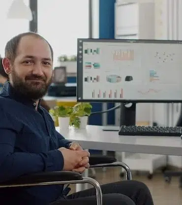 Man in a wheelchair smiling at the camera in a modern office with colleagues working in the background.