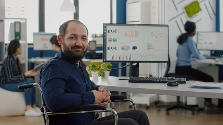 Man in a wheelchair smiling at the camera in a modern office with colleagues working in the background.