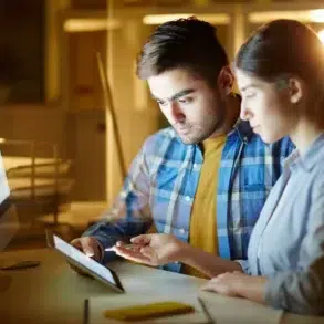 Two people look at a tablet in a dimly lit office with a computer screen in the background.