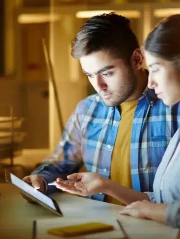 Two people look at a tablet in a dimly lit office with a computer screen in the background.