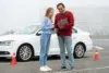 A driving instructor and a student review notes beside a parked white car. Orange traffic cones are in the background. They appear focused and engaged.