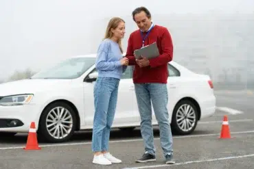 A driving instructor and a student review notes beside a parked white car. Orange traffic cones are in the background. They appear focused and engaged.