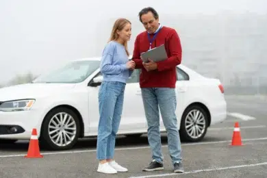 A driving instructor and a student review notes beside a parked white car. Orange traffic cones are in the background. They appear focused and engaged.