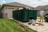 A man stands next to a large green dumpster filled with construction debris in front of a garage.