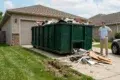 A man stands next to a large green dumpster filled with construction debris in front of a garage.