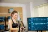 Woman exercising on an elliptical trainer with electrodes attached to her chest, monitored by multiple screens displaying vital signs and ECG readings.