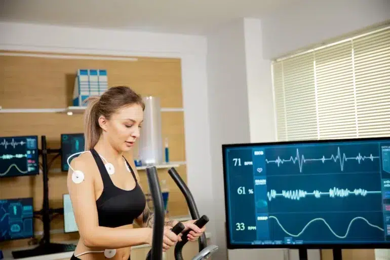 Woman exercising on an elliptical trainer with electrodes attached to her chest, monitored by multiple screens displaying vital signs and ECG readings.