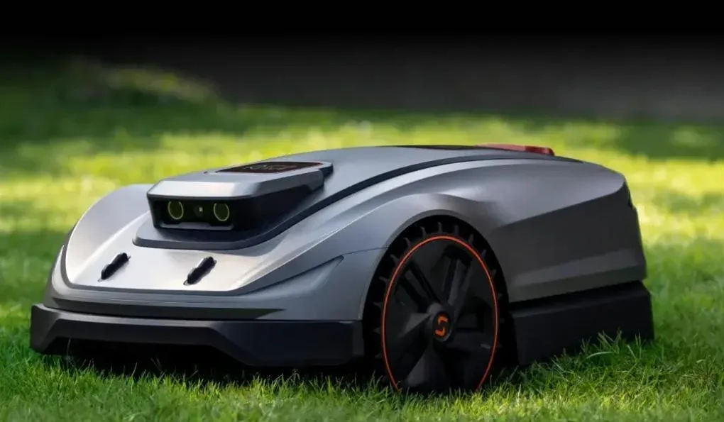 Sleek robotic lawn mower with sensors and orange-accented wheels rests on green grass.