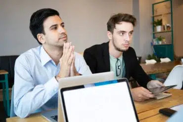 Two men in business attire at a table with laptops and a tablet.