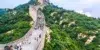 Tourists walk on the Great Wall of China winding through a lush green, hilly landscape under a cloudy sky.