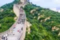 Tourists walk on the Great Wall of China winding through a lush green, hilly landscape under a cloudy sky.
