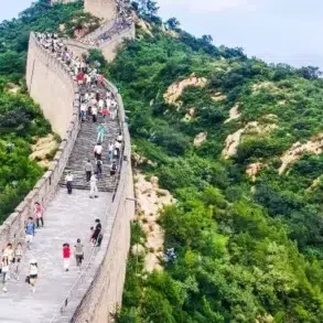 Tourists walk on the Great Wall of China winding through a lush green, hilly landscape under a cloudy sky.