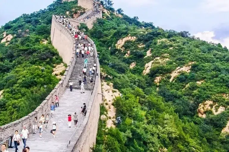 Tourists walk on the Great Wall of China winding through a lush green, hilly landscape under a cloudy sky.