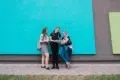 Three casually dressed teenagers with backpacks stand and talk in front of a bright blue wall.