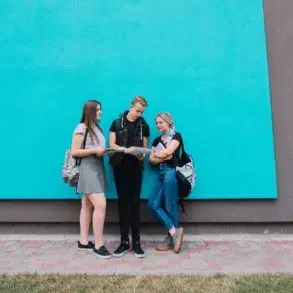 Three casually dressed teenagers with backpacks stand and talk in front of a bright blue wall.