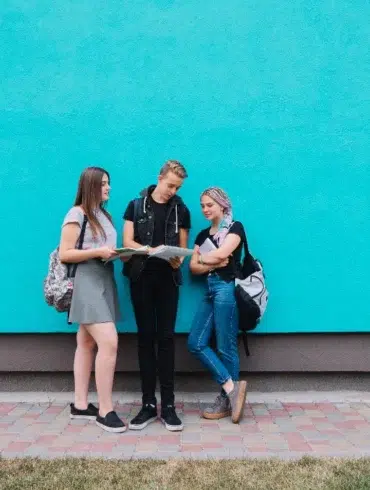 Three casually dressed teenagers with backpacks stand and talk in front of a bright blue wall.