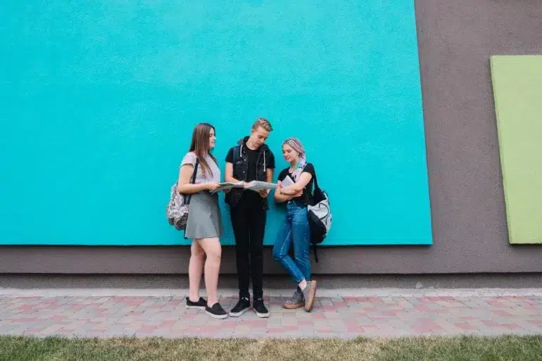Three casually dressed teenagers with backpacks stand and talk in front of a bright blue wall.