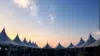A row of white pointed tents is silhouetted against a vibrant sunset sky, creating a serene and festive atmosphere. People gather underneath.
