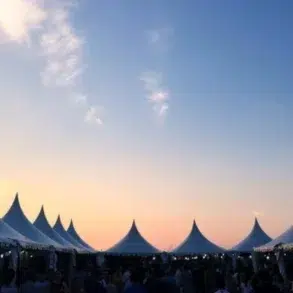 A row of white pointed tents is silhouetted against a vibrant sunset sky, creating a serene and festive atmosphere. People gather underneath.