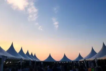 A row of white pointed tents is silhouetted against a vibrant sunset sky, creating a serene and festive atmosphere. People gather underneath.