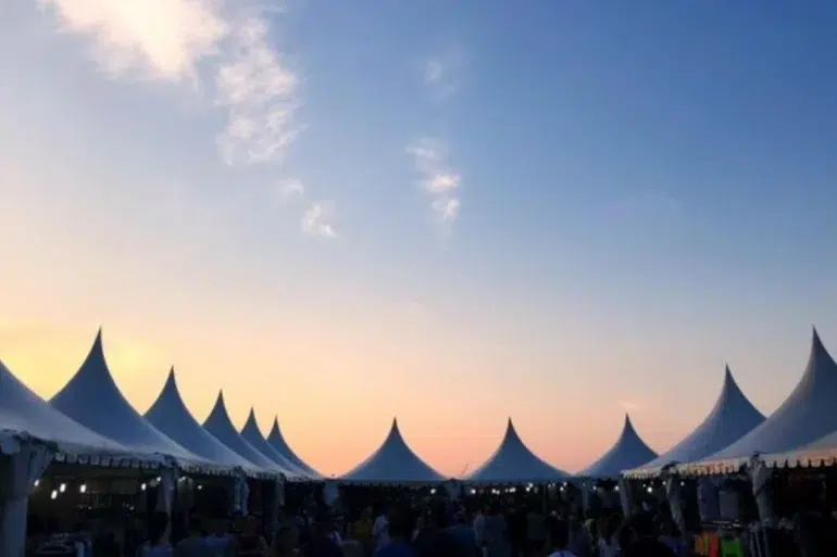 A row of white pointed tents is silhouetted against a vibrant sunset sky, creating a serene and festive atmosphere. People gather underneath.