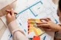 A person reviews architectural blueprints with a color palette in hand, selecting shades of yellow, orange, and red. The scene conveys creativity.