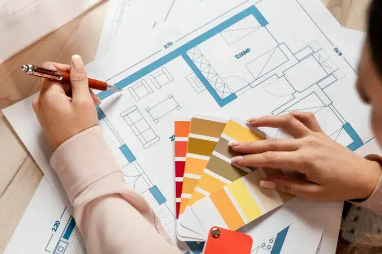 A person reviews architectural blueprints with a color palette in hand, selecting shades of yellow, orange, and red. The scene conveys creativity.