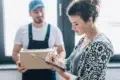 A delivery person holds a box while a woman signs a document on a clipboard.